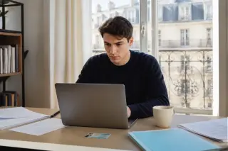 Un jeune adulte prépare sa déclaration d'activité sur un ordinateur portable, entouré de documents administratifs dans un appartement lumineux.